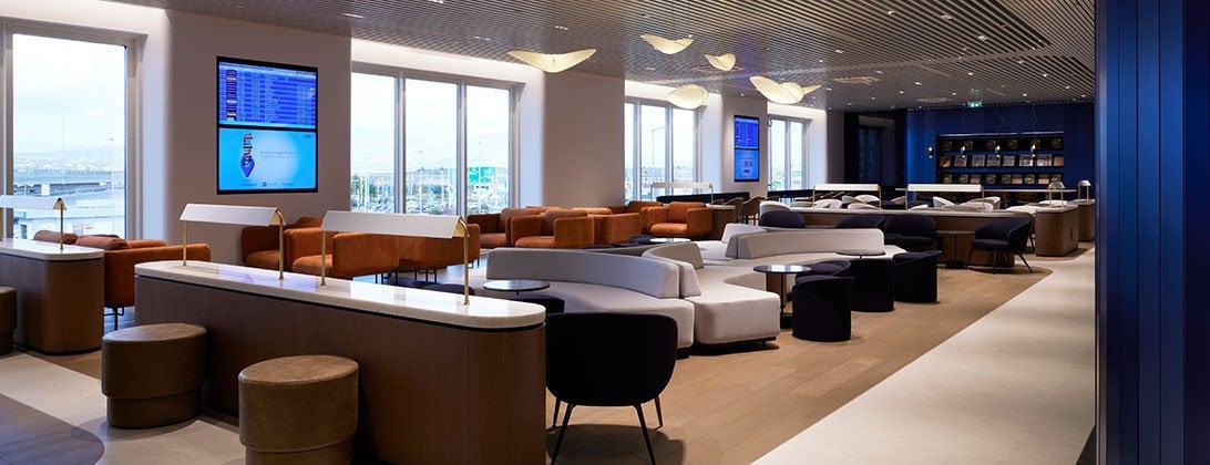 The Hall B Aegean Lounge at Athens airport showcases a modern interior with panoramic window views of the airport. The lounge has a variety of seating options such as tan leather chairs, white sofas and round stools.