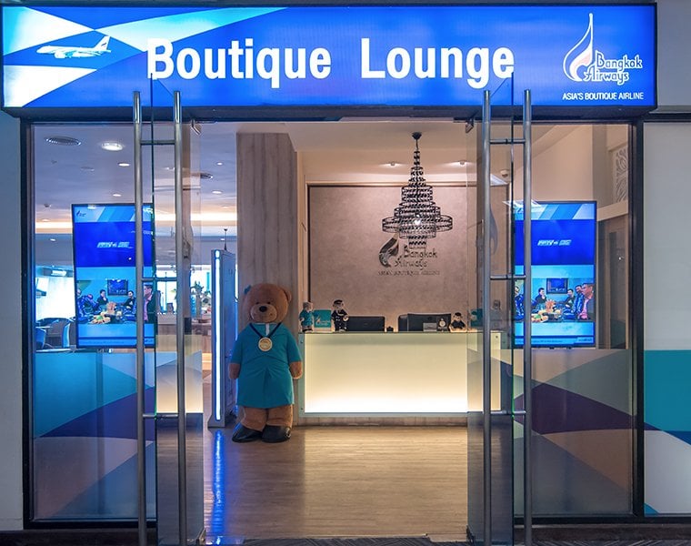 Entrance to the Bangkok Airways Boutique Lounge at Chiang Mai International Airport (CNX), featuring a bright blue sign, glass doors, a welcoming reception desk, and a large teddy bear mascot dressed in airline uniform under a chandelier wall decal.