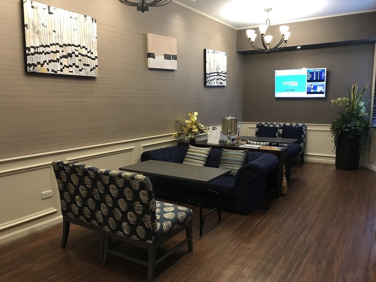 Cozy seating area inside the Blue Ribbon Club Lounge at Chiang Mai International Airport (CNX), featuring navy upholstered sofas with patterned cushions, a wall-mounted flight information screen, and modern abstract artwork.