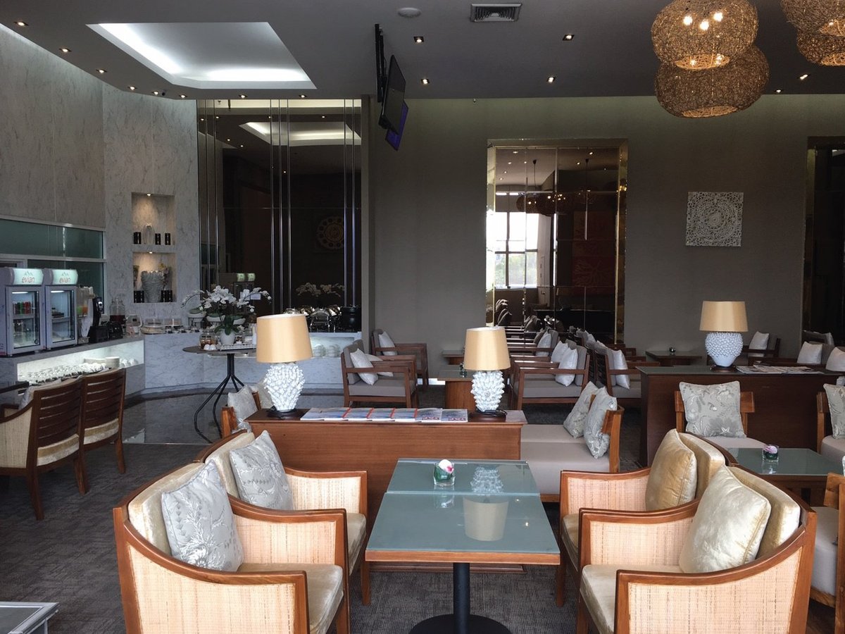Interior view of The Coral Executive Lounge at Chiang Mai International Airport (CNX) International Terminal, showcasing elegant seating with rattan and upholstered armchairs, glass-top tables, and marble buffet counters.
