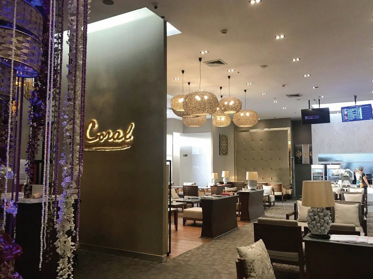 Entrance and interior of The Coral Executive Lounge at Chiang Mai International Airport (CNX) International Terminal, featuring warm taupe walls, elegant pendant lighting, and cream-upholstered seating with dark wood frames.
