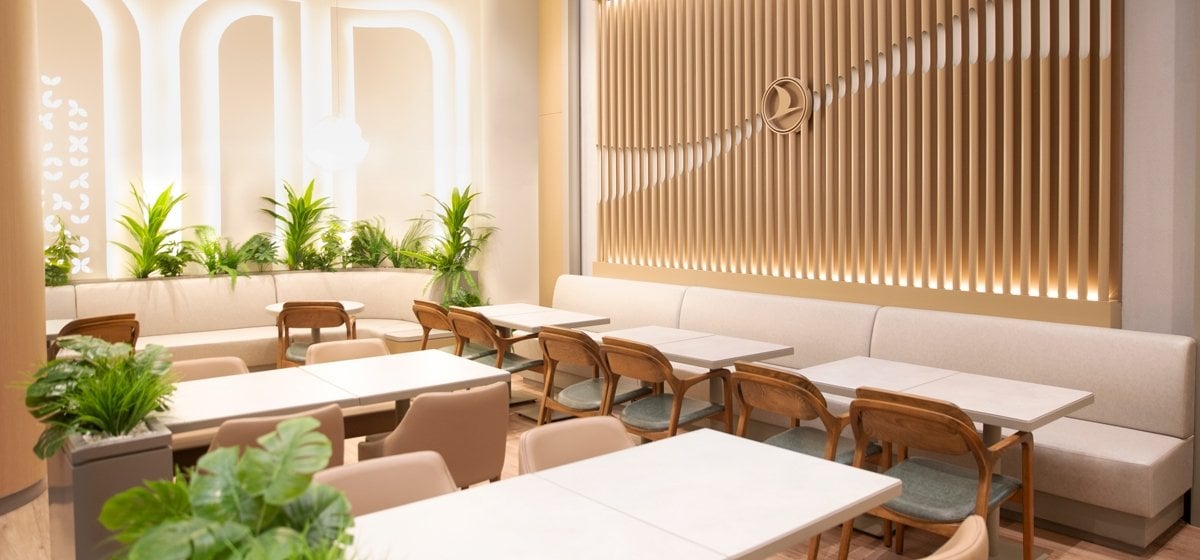Bright and elegant dining area inside the Turkish Airlines CIP Lounge at Çukurova International Airport, featuring soft lighting, modern booth seating, greenery accents, and the airline’s signature logo on a wood-paneled wall.