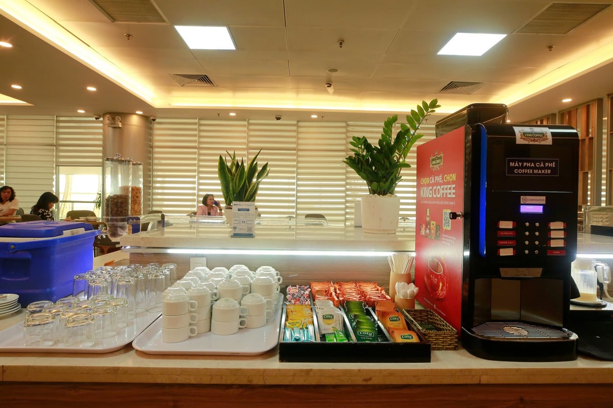 Beverage station at Vietnam Airlines Lotus Lounge in Da Nang Airport featuring a coffee machine, assorted tea and coffee sachets, stacked cups and glasses, and a clean, modern counter with indoor plants.