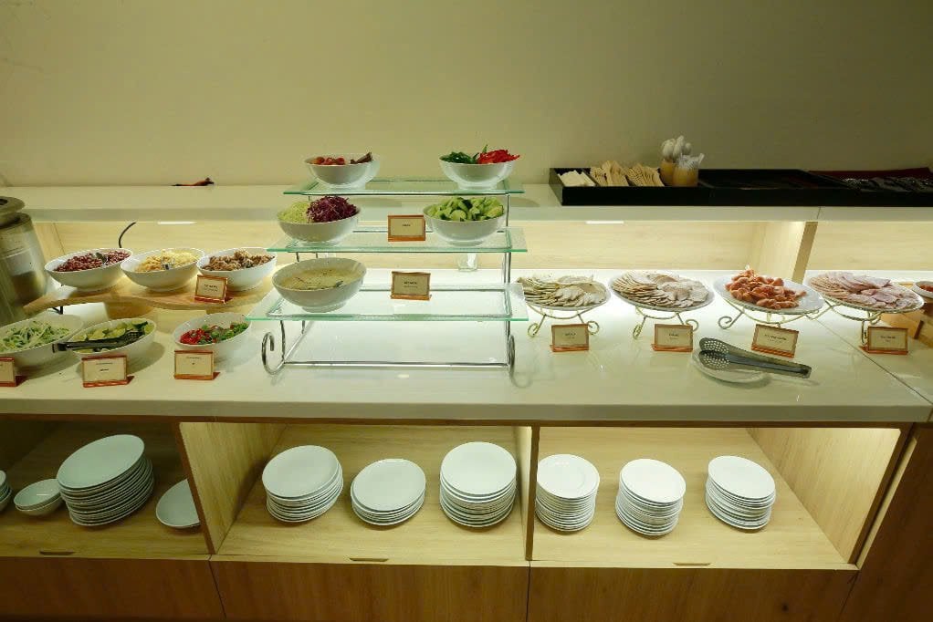 Buffet counter at C2 Business Class Lounge in Da Nang Airport displaying cold cuts, fresh salad ingredients, dressings, and condiments, with neatly stacked plates and bowls beneath the counter.