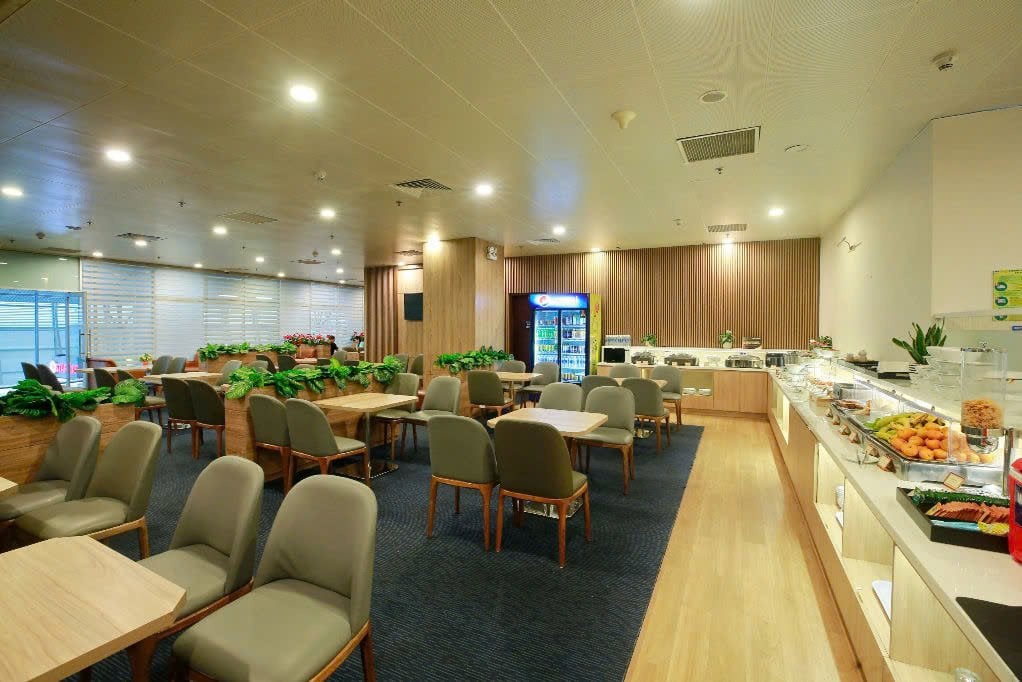 Interior of Business Class Lounge C2 at Da Nang Airport featuring dining tables, buffet with fresh fruit and snacks, beverage fridge, and relaxed seating in a modern, wood-accented setting.