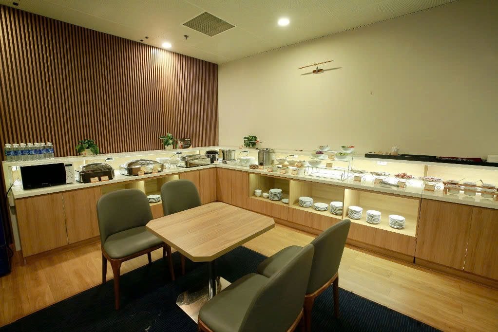 Dining area at Business Class Lounge C2 in Da Nang Airport featuring a hot and cold buffet spread, modern wood-paneled counters, stacked plates, and a small seating table with two chairs.