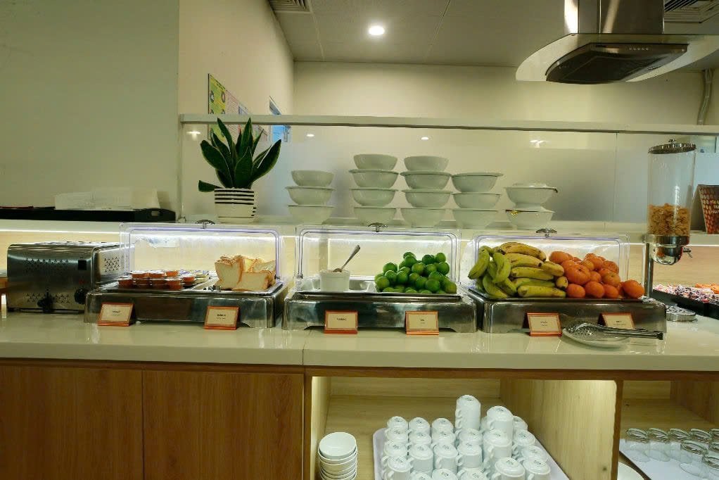 Breakfast and fruit counter at C2 Business Class Lounge in Da Nang Airport, featuring sliced bread, jam, limes, bananas, oranges, cereal dispenser, and neatly stacked bowls and mugs.