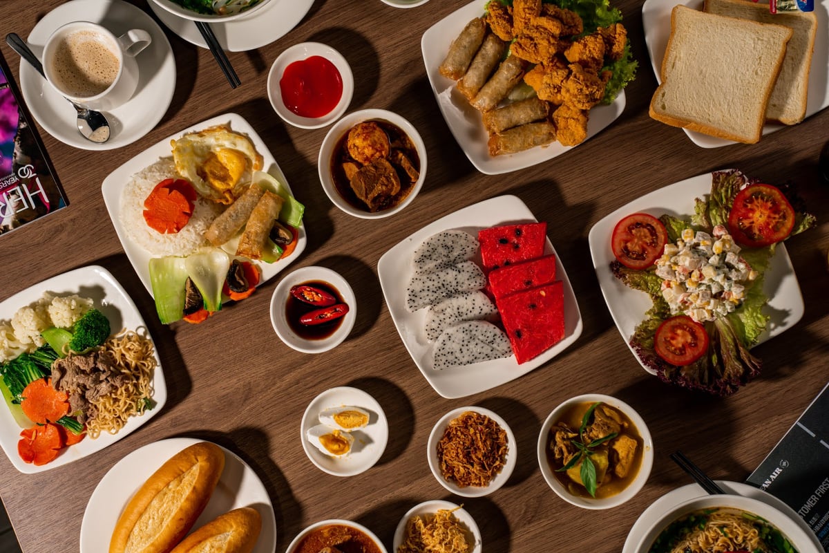 Assorted Vietnamese and international dishes served at CIP Orchid Lounge 1 in Da Nang Airport Terminal 2, including rice with egg and spring rolls, banh mi, noodle stir-fry, tropical fruits, fresh salads, and traditional soups on a wooden table.