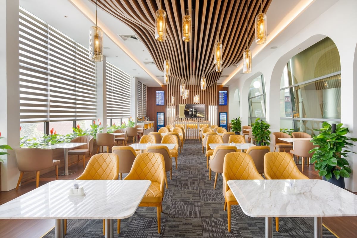 Spacious seating area inside Vietcombank Priority Lounge at Da Nang Airport, featuring modern yellow and beige armchairs, white marble-top tables, lush green plants, and an eye-catching wave-style wooden ceiling with pendant lights.