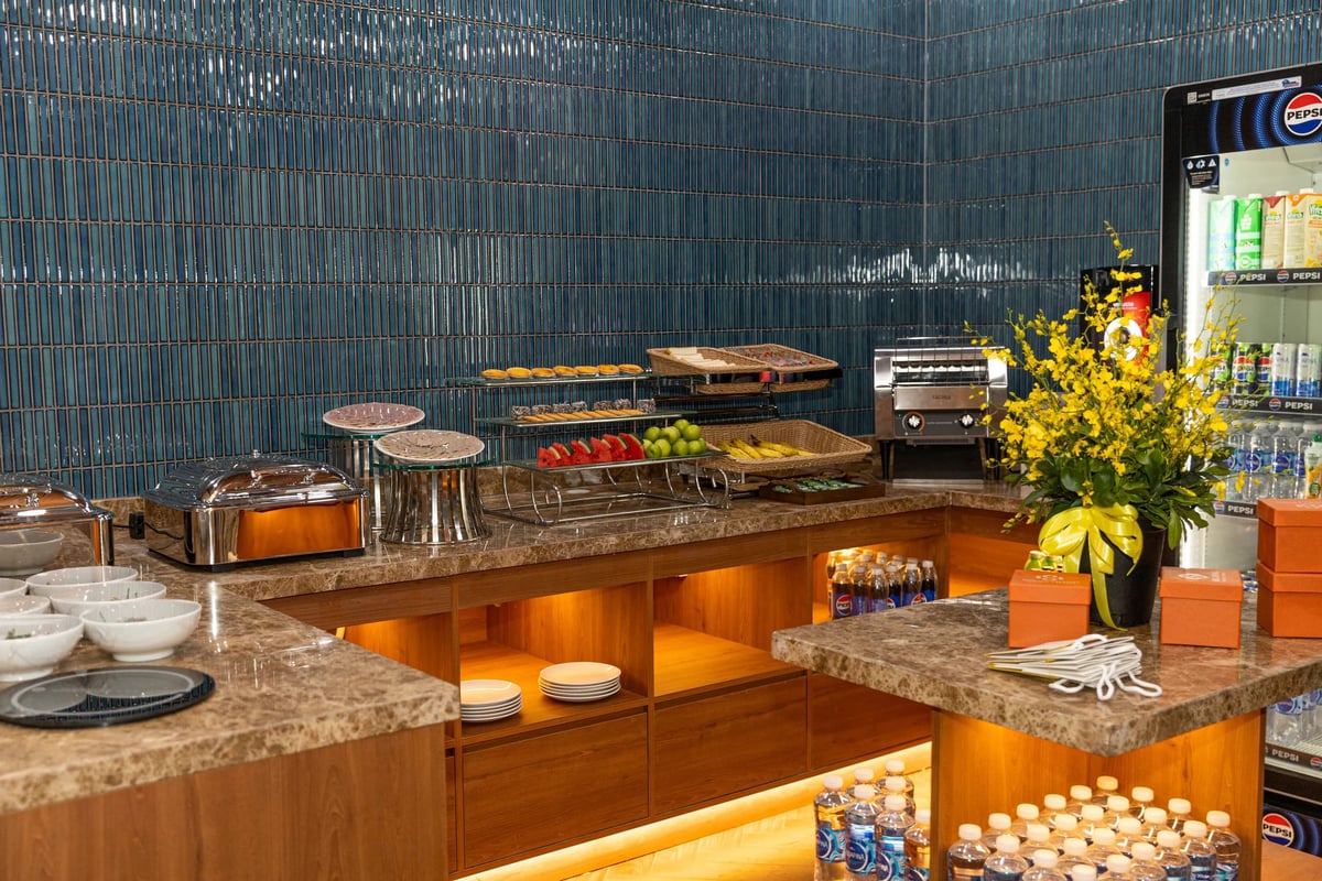 Buffet spread at SH Premium Lounge in Da Nang International Airport featuring fresh fruits, pastries, hot dishes, bottled drinks, and snacks, set against a modern blue tile wall and marble countertop.