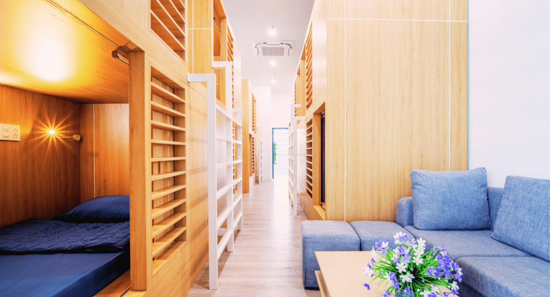 Modern sleep pod corridor at Da Nang International Airport’s Sleepod facility, featuring wooden enclosed bunk-style pods with navy bedding, warm ambient lighting, and a cozy lounge area with a blue sectional sofa and floral centerpiece.