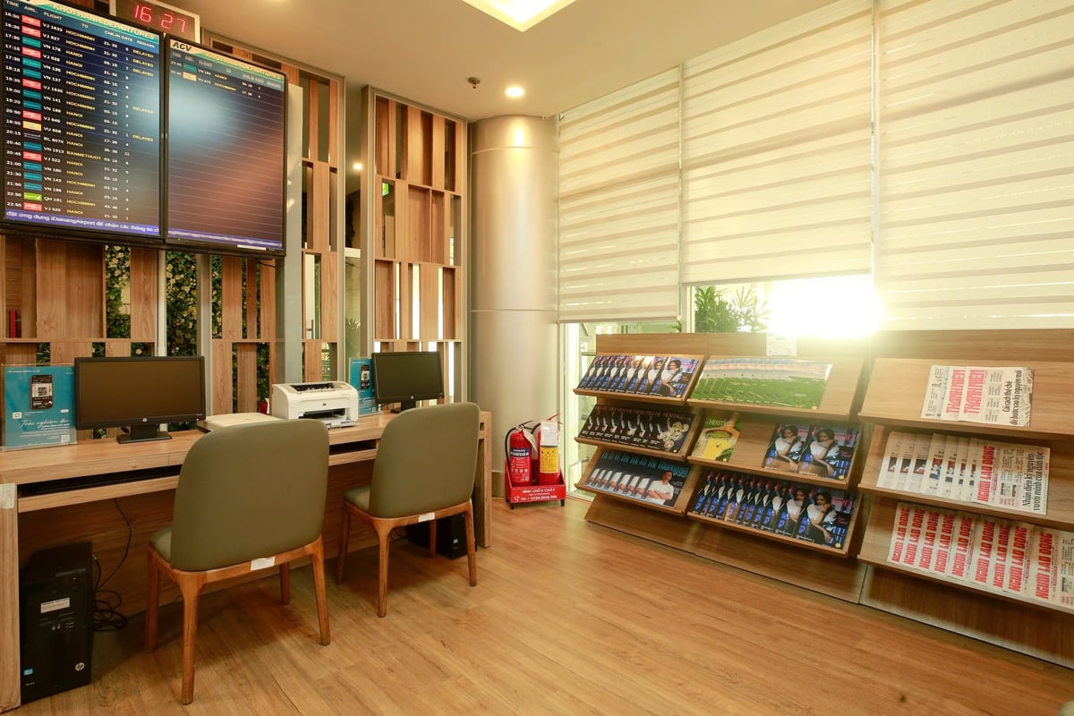 Workstation and reading area inside Vietnam Airlines Lotus Lounge at Da Nang Airport, featuring desktop computers, a printer, flight information screens, and a well-stocked magazine rack by large sunlit windows.