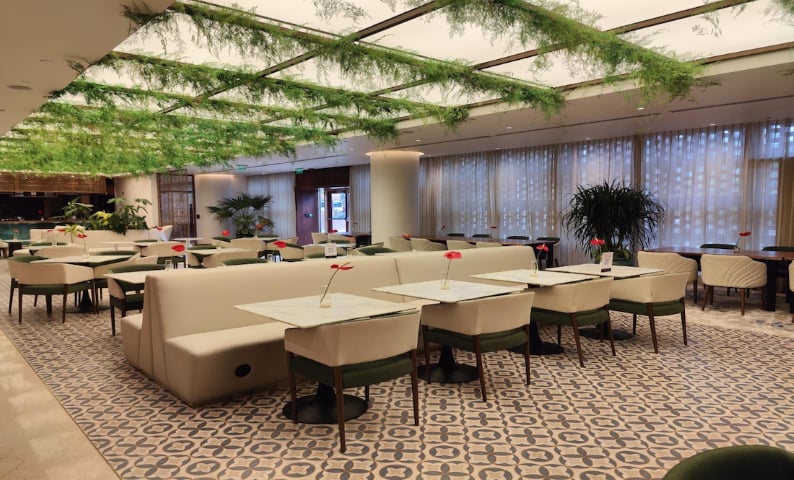 Interior view of Arrival Lounge, Bangalore Airport. The space features a modern design with neutral tones, including beige seating and tables arranged in a neat layout. The ceiling is adorned with greenery, creating a natural and fresh atmosphere.