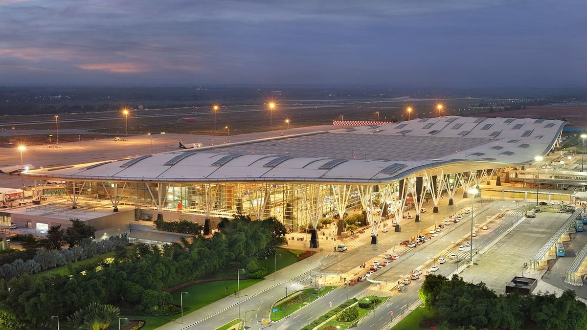 Aerial view of Kempegowda International Airport in Bangalore during twilight. The image captures the sprawling airport structure with its distinctive, futuristic roof design, illuminated by both the setting sun and airport lighting.