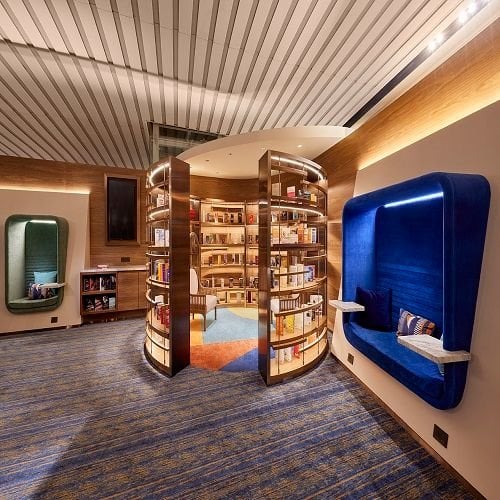 This photo features a cozy reading nook within an airport lounge. The space is designed with modern aesthetics, featuring a slatted wood ceiling and vibrant carpet in shades of blue, orange, and beige.