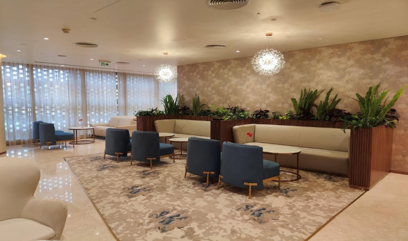 Vip section of the arrival lounge at BLR airport.The space includes blue upholstered chairs and beige sofas arranged around small wooden tables, each adorned with a single red flower in a vase.