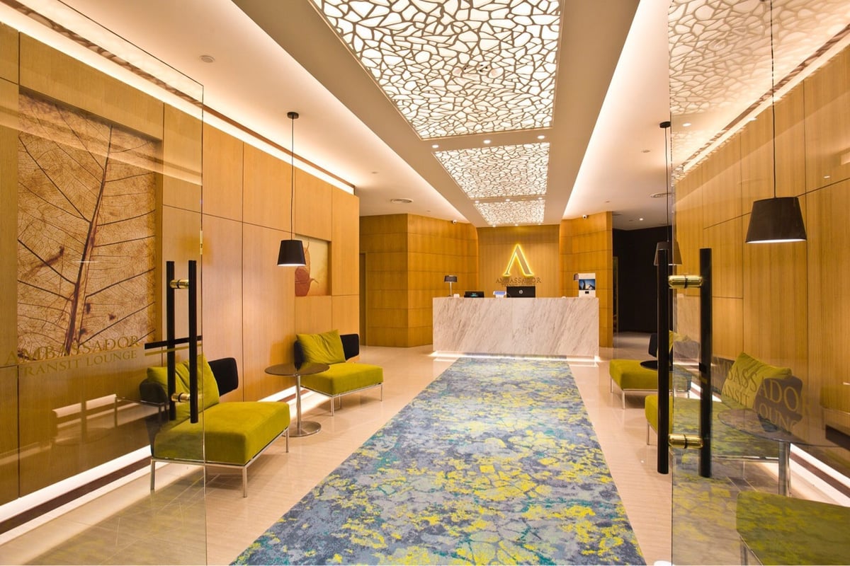 Reception area of Ambassador Transit Lounge at Changi Airport Terminal 2, featuring modern wood paneling, a marble front desk, decorative ceiling lighting, lime green seating, and a patterned blue and yellow carpet.