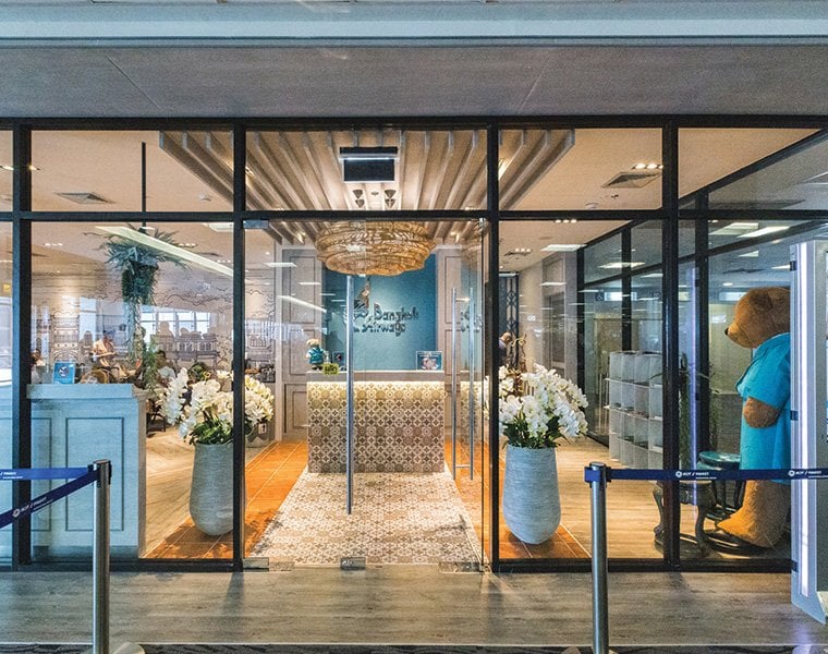 This image features the entrance to the Bangkok Airways Boutique Lounge at Phuket Airport. The front of the lounge is visible through clear glass doors, showcasing a stylish and welcoming interior.