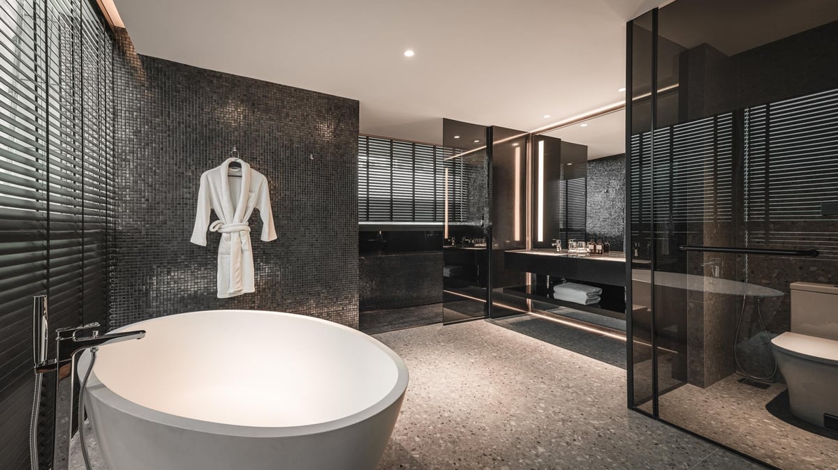 Luxurious bathroom at Capsule Transit MAX in KLIA Terminal 2, featuring a deep standalone bathtub, a hanging white robe, dark mosaic tiled walls, a spacious vanity area with a large mirror, modern fixtures, and sleek lighting accents.