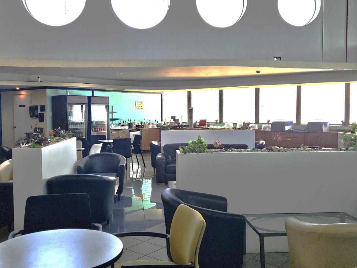 Seating and dining area of the BGI Airlines Executive Lounge featuring leather black armchairs, yellow dining chairs and round tables, and a buffet and beverage station in the background.
