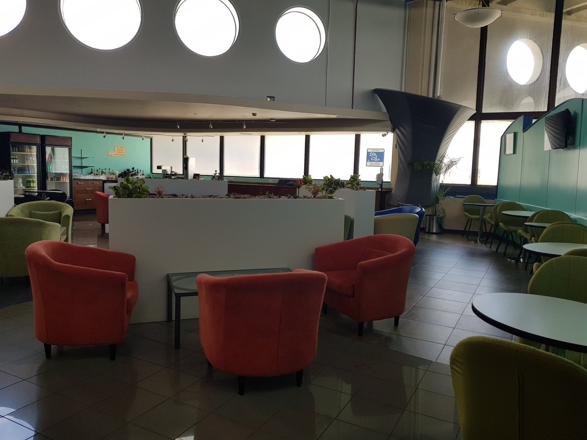 Dining and lounge area inside the Airlines Executive Lounge at Grantley Adams International Airport, featuring colorful armchairs, beverage station, and modern circular windows.