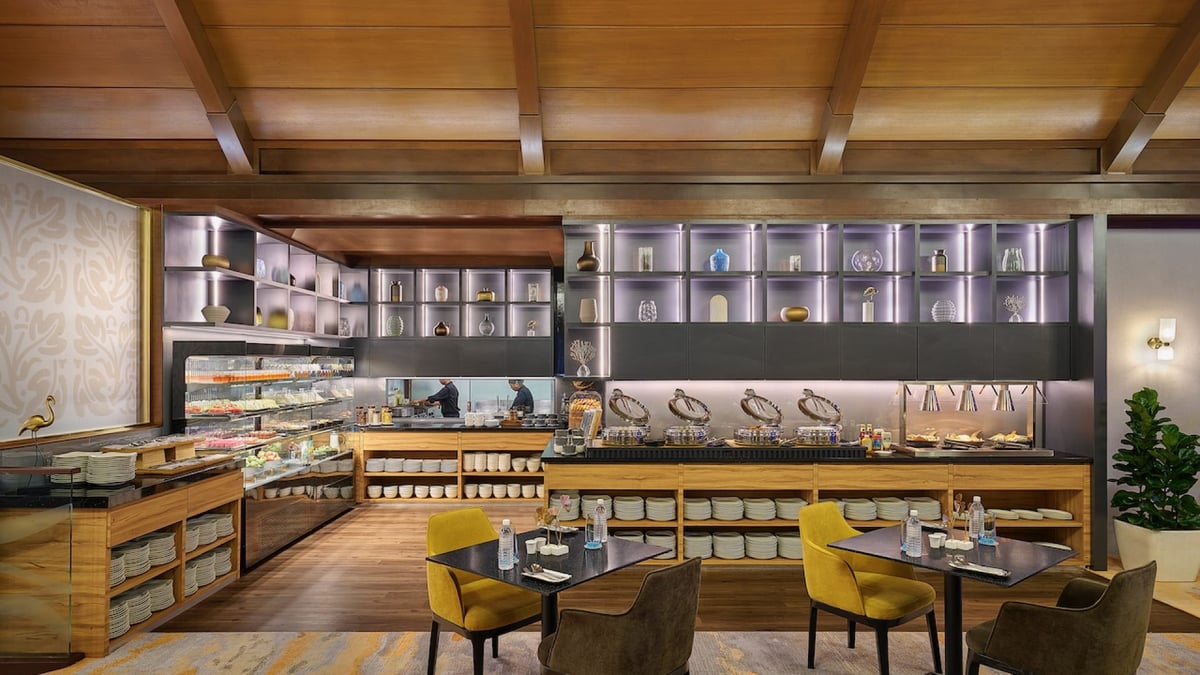 Buffet area at the Sky Suite Satellite Terminal Kuala Lumpur Airport, featuring a stylish food counter with a variety of dishes, a refrigerated display of cold items, and nearby dining tables that have mustard and brown chairs.