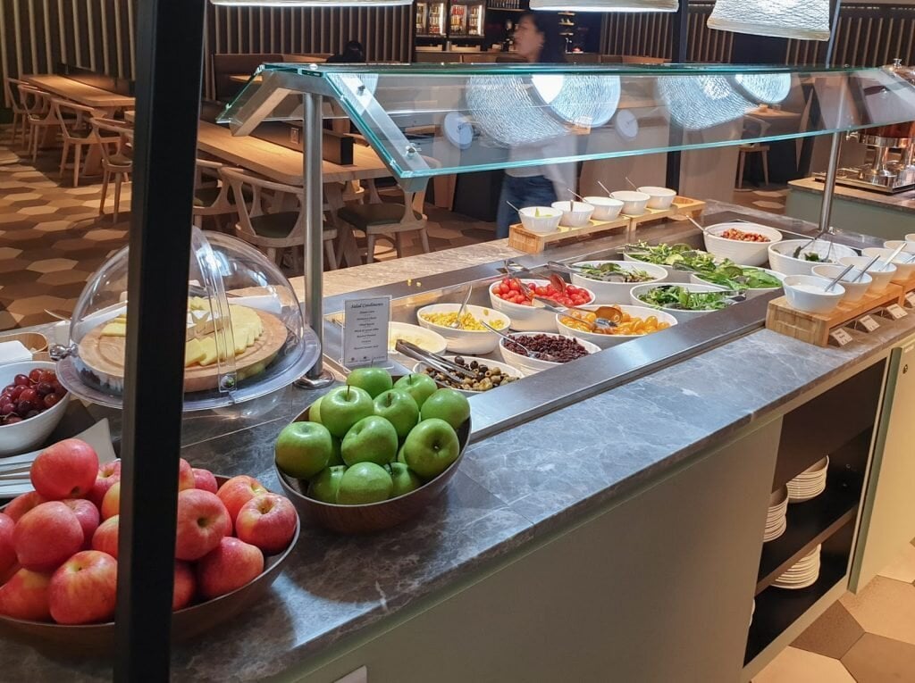 Buffet salad and fresh fruit counter at Blossom Lounge Changi Airport Terminal 4 with apples, grapes, and assorted vegetables.