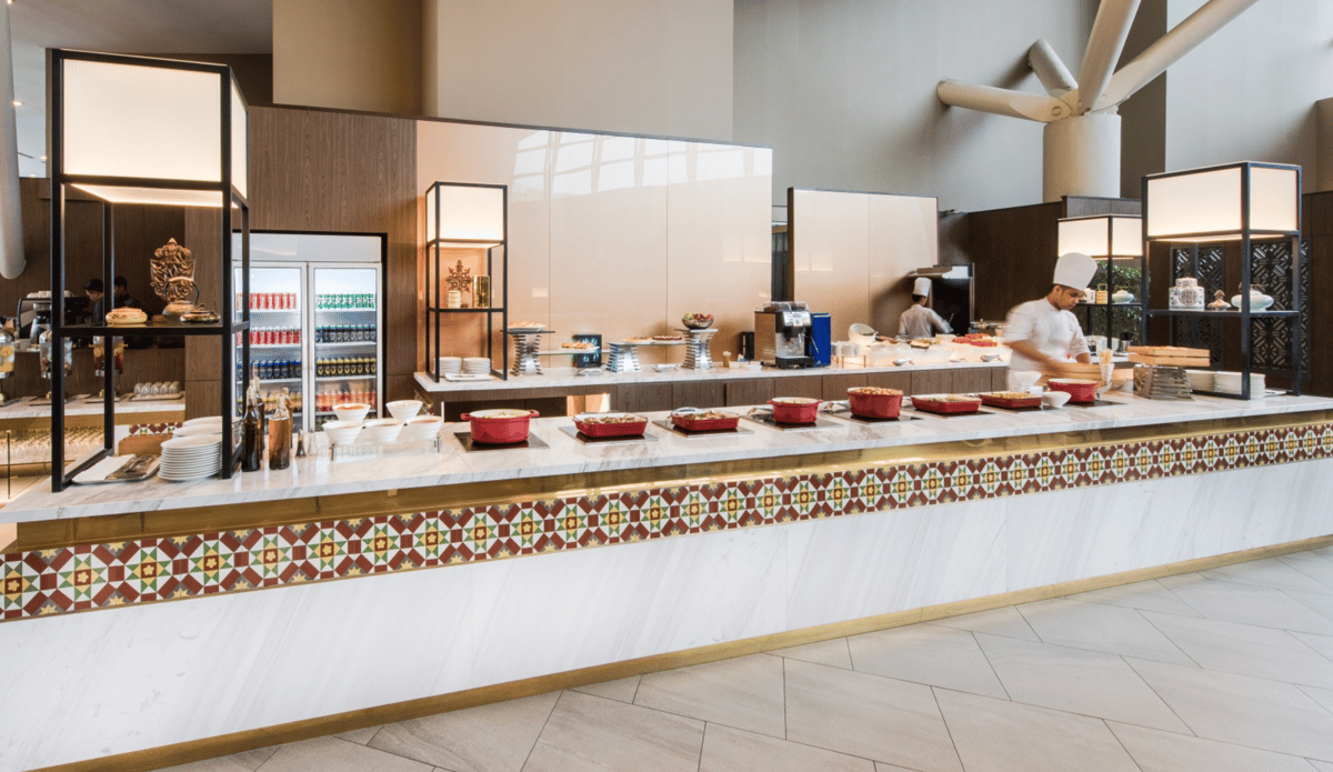 Buffet counter at Malaysia Airlines’ Regional Golden Lounge, KLIA Terminal 1, featuring a selection of hot dishes in red pots, a drinks fridge, modern food displays, and chefs preparing fresh items in an open kitchen setting.