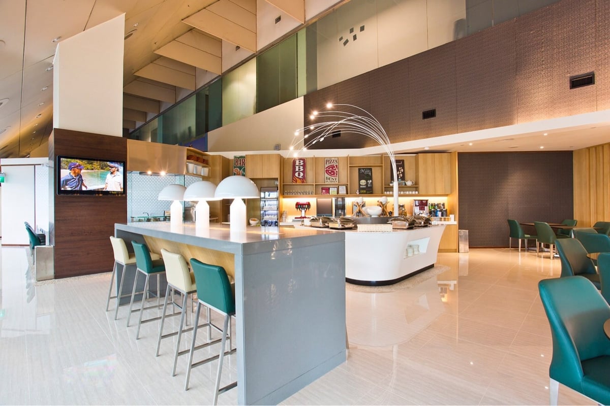 Buffet area inside Ambassador Transit Lounge at Changi Airport Terminal 2, featuring a sleek modern counter with built-in food warmers, bright overhead lighting, and teal dining chairs, offering a variety of hot dishes and beverages for travelers.