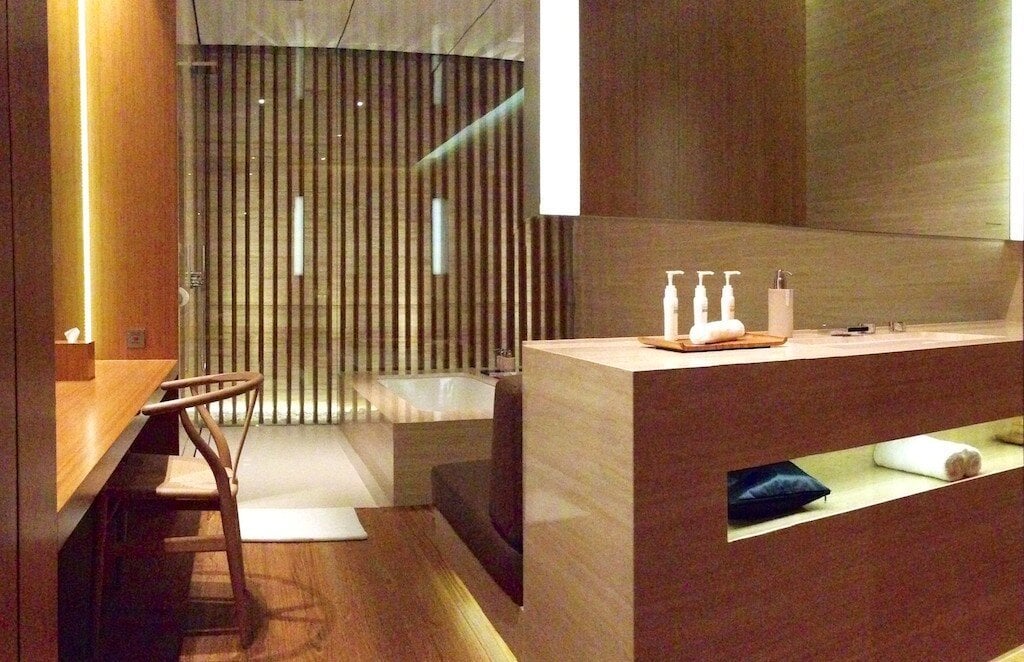 Interior of a Cabana suite at The Wing First Class Lounge in Hong Kong International Airport, featuring a deep soaking tub behind slatted wood dividers, a spacious vanity with toiletries, a wooden desk and chair, and ambient lighting.