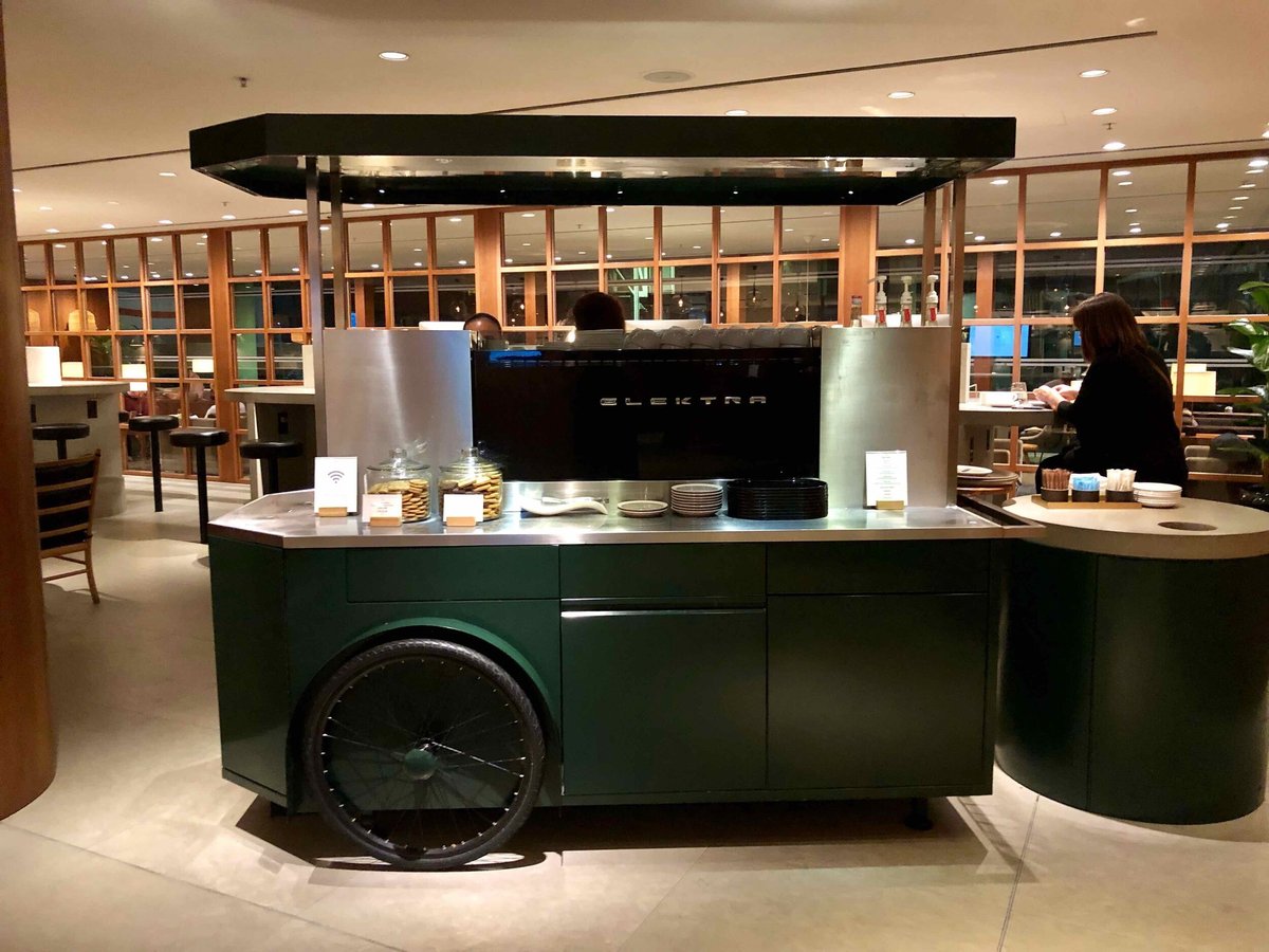 A sleek modern coffee cart at the Pier Business Class Cathay Pacific Lounge HKG equipped with an Elektra espresso machine, jars of cookies, stacked plates and a station for coffee condiments.