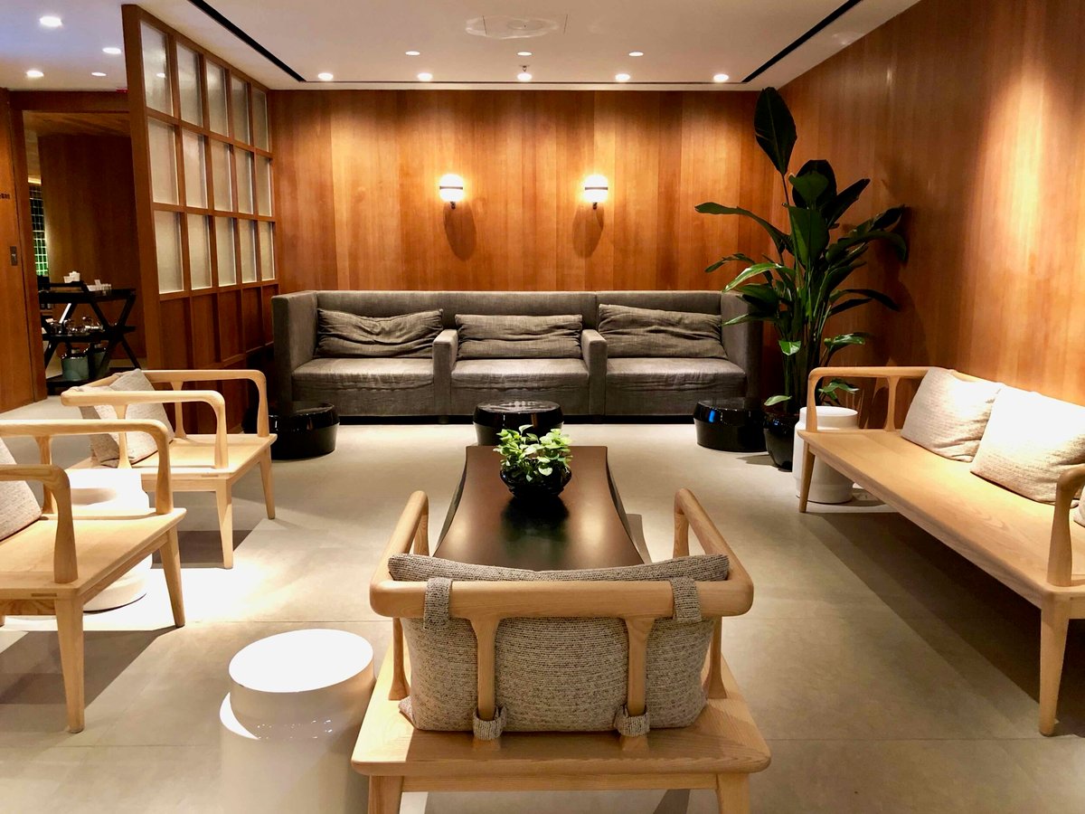 Warmly lit seating area in Cathay Pacific's The Pier Business Class Lounge at Hong Kong Airport featuring a grey sofa, and light wooden seats with a room partition to the left.