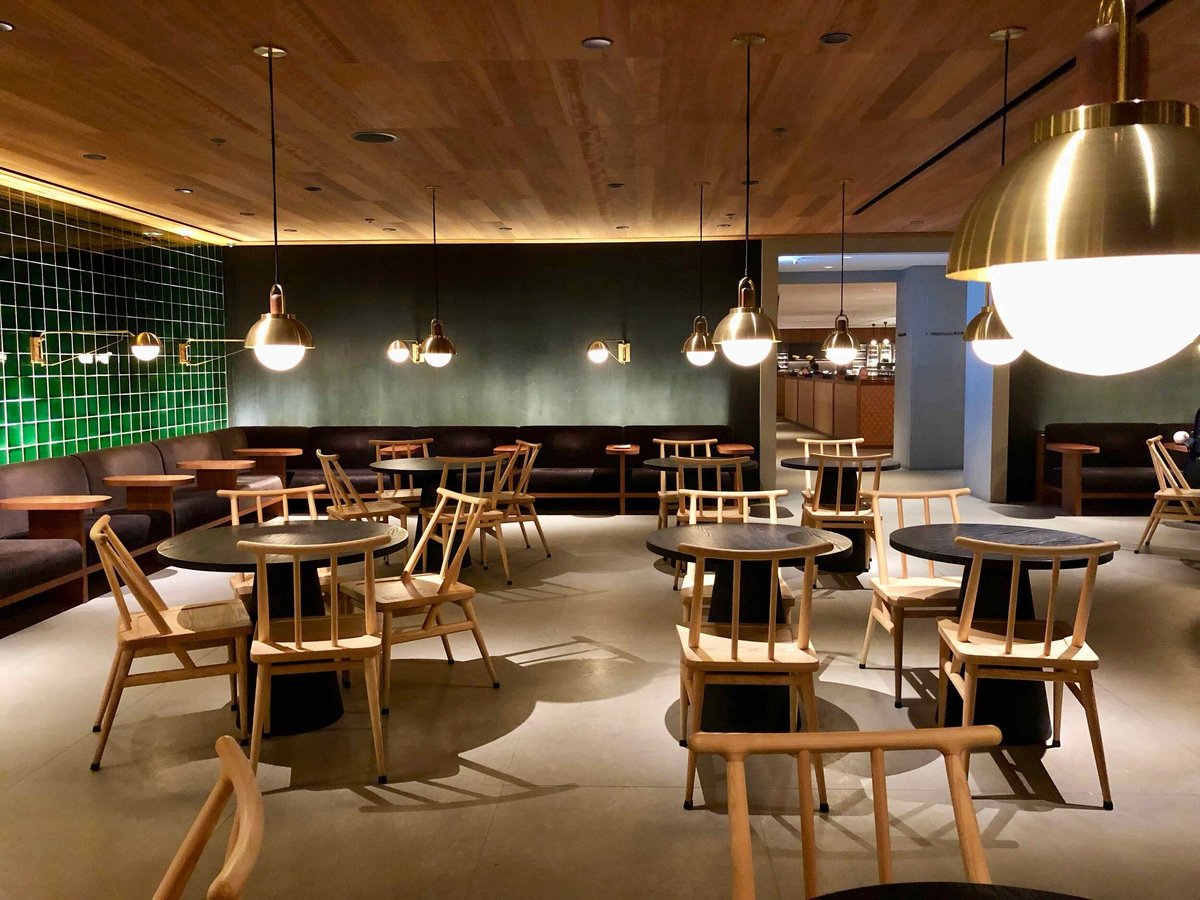 The Teahouse seating area at the Business Class Pier Cathay Pacific Lounge HKG featuring round black tables and light wooden chairs, lighting with brass accents, a dark green tiled wall, and built in cushioned bench seating for a cozy atmosphere.
