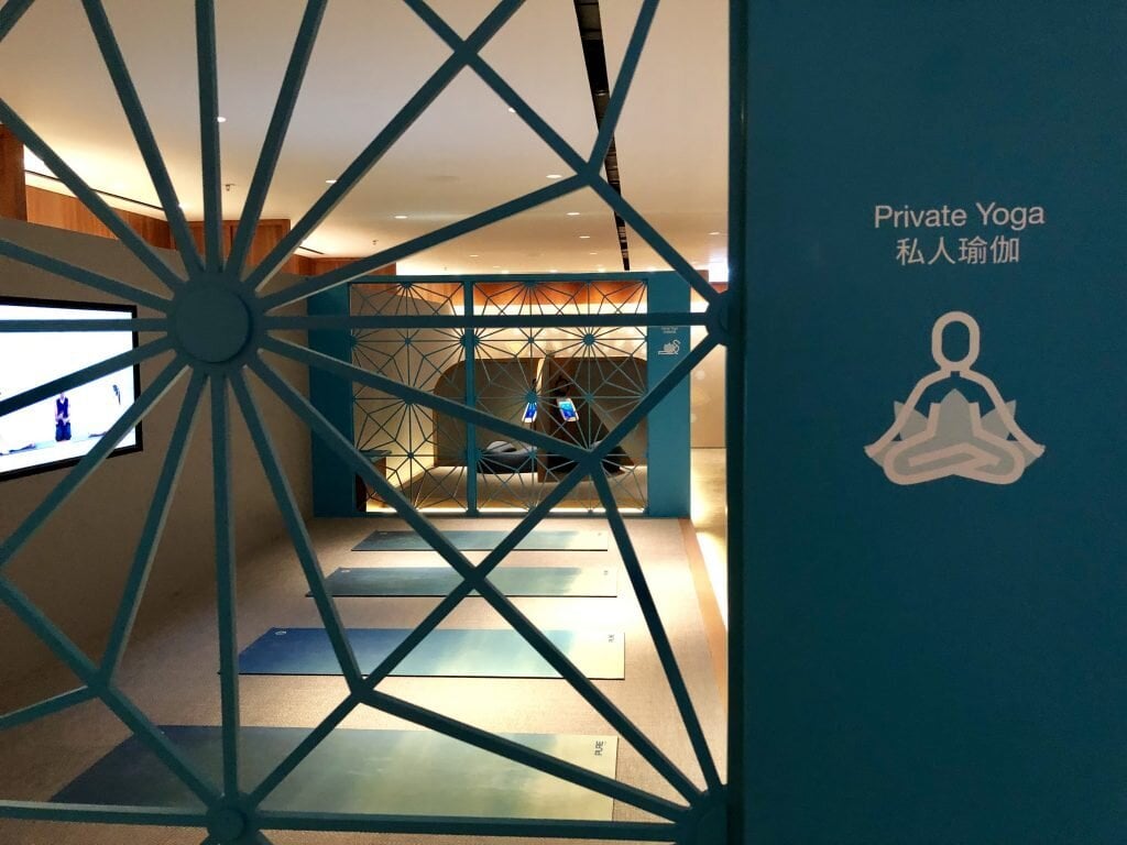 The Private Yoga area inside The Sanctuary at The Pier Business Class Lounge in Hong Kong International Airport, featuring blue yoga mats aligned on a carpeted floor, decorative lattice partitions, and a screen displaying guided yoga instructions.