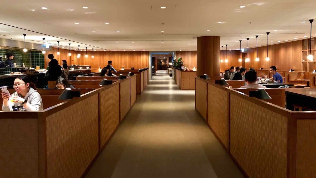 Wooden dining booths, a long central walkway and chefs preparing freshly made dishes behind a sleek counter with green tiled walls at the Pier Business Lounge at Hong Kong International Airport.