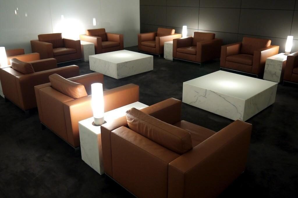 A quiet seating area inside the Cathay Pacific The Wing Business Class Lounge, featuring a symmetrical layout of brown leather armchairs surrounding white marble tables, set against a dark carpet and minimalist wall panels.