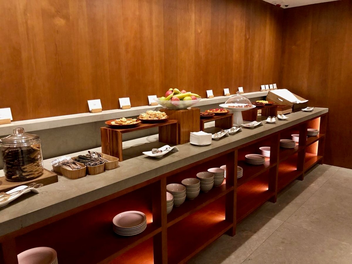 The Pantry at The Pier First Class Lounge in Hong Kong International Airport, displaying a selection of light snacks including cookies, fresh fruit, pastries, and cakes, with plates, bowls, and cutlery neatly organized on open shelving.