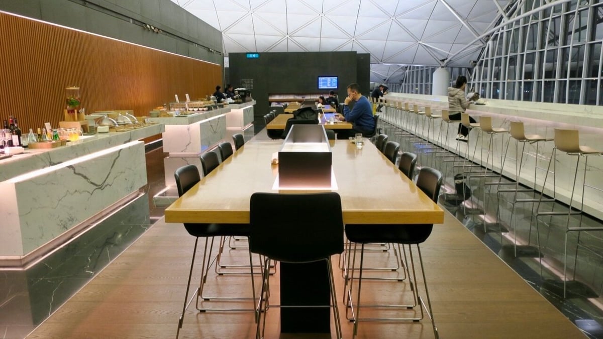 Dining area in The Wing First Class Lounge at Hong Kong International Airport, featuring long communal tables with black chairs, a marble buffet counter with food and beverages, and a high bar-style counter.