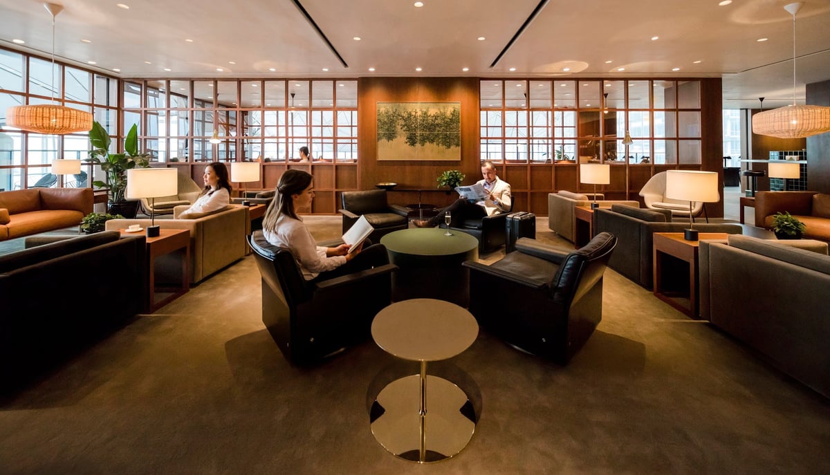 The seating area at Cathay Pacific’s The Deck lounge in Hong Kong International Airport, featuring modern armchairs, soft lighting from table lamps, wooden partitions, earth-toned décor, and passengers reading in a relaxed, quiet environment