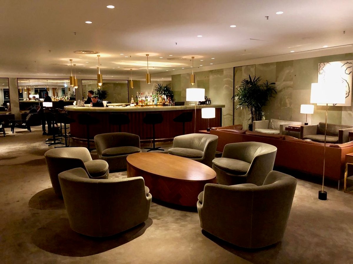 The bar area at The Pier First Class Lounge in Hong Kong International Airport, featuring a circular marble-topped bar with bar stools, plush lounge chairs arranged around a wooden coffee table, soft ambient lighting, and elegant decor.