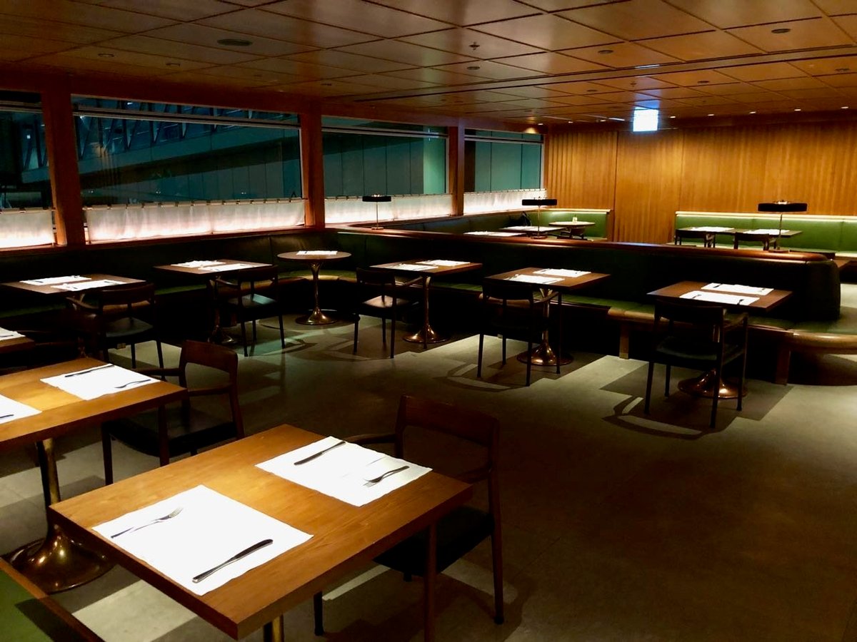 The Dining Room at The Pier First Class Lounge in Hong Kong International Airport, featuring neatly set wooden tables with brass bases, green upholstered banquette seating, soft overhead lighting, and a calm, refined ambiance.
