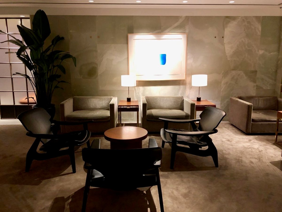 A quiet seating area at The Pier First Class Lounge in Hong Kong International Airport, featuring neutral-toned armchairs and modern black lounge chairs arranged around a circular wooden table.