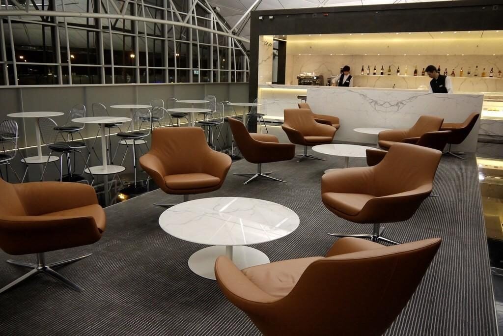 The Long Bar Area in The Wing Business Class Lounge at HKG Airport, featuring a sleek white marble bar staffed with bartenders, modern leather armchairs surrounding whtie tables and cafe style seating near the window overlooking the Terminal.