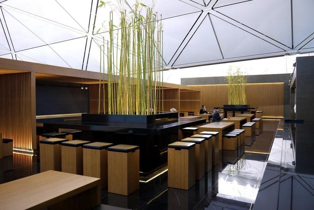 Modern seating area inside the Cathay Pacific The Wing Business Class Lounge at HKG, featuring minimalist wooden stools and tables surrounding a central island with tall bamboo plants.