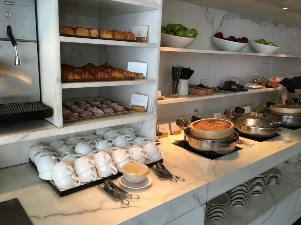 Buffet area in the wing featuring stacked rows of white coffee mugs, pastries such a croissants in storage section overhead, and hot meals along the rest of the counter.