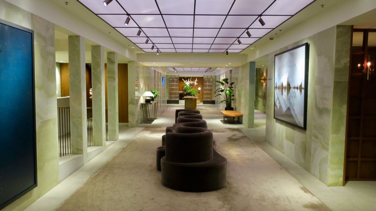 The entrance corridor of The Pier First Class Lounge at Hong Kong International Airport, featuring polished green onyx walls, modern art, soft carpeted flooring, ambient lighting, and curved velvet seating with plants and elegant furnishings.