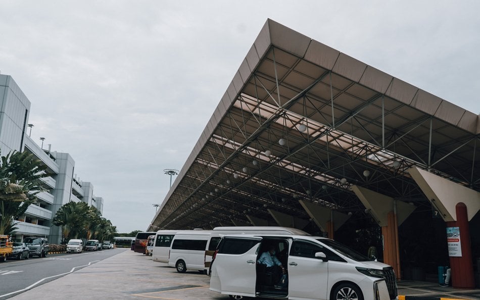 Vehicle pick-up and drop-off area at Changi Airport Terminal 2, featuring a covered bay with a large slanted roof structure, multiple vans and cars, and the airport’s modern exterior buildings in the background.