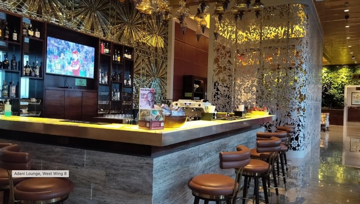 Bar at the Terminal 2 Adani Lounge West Wing at Mumbai Airport, featuring lace patterned partitions that separate the bar from the seating area. The bar has several high stools and an array of utensils on the counter, behind the bar is alcohol.