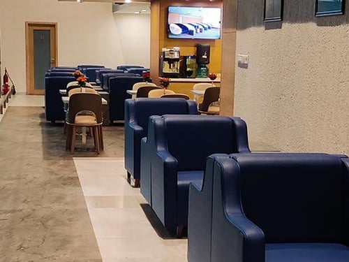 Blue armchairs at the East wing aviserv Lounge at t2 Mumbai Airport. There are some dining options to the back of the room and what appears to be a beverage stations and television above the station.