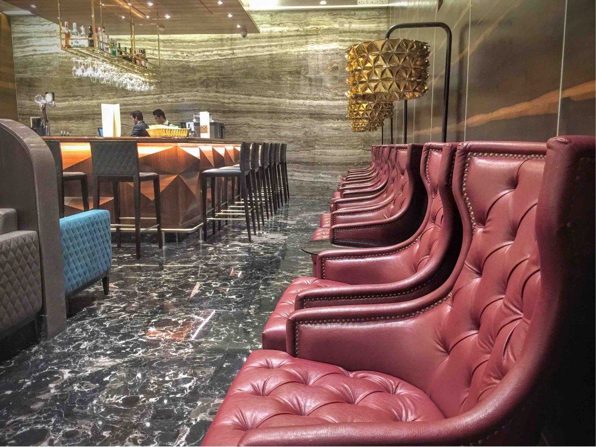 Leather ruby red armchairs against a back wall of the T2 Travel Club Lounge at Mumbai Airport. There are golden light fixtures behind the chairs and a bar to the left with attendants seen working.